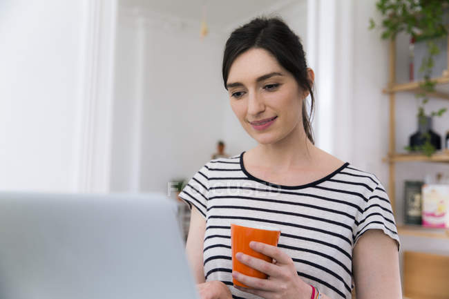 Beautiful caucasian brunette woman with cup sitting at home and using laptop — Stock Photo