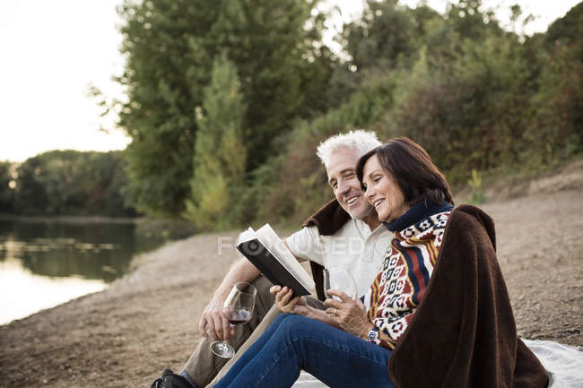 Feliz pareja de ancianos con vino y libro en un lago - foto de stock