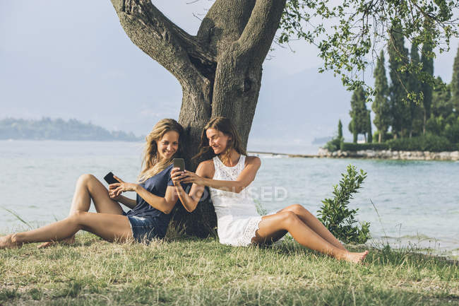 Italien, Gardasee, zwei junge Frauen lehnen mit Mobiltelefonen an einem Baum — Stockfoto