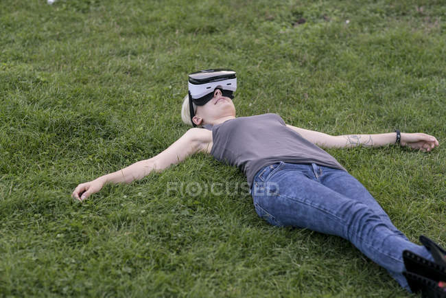 Jeune femme allongée sur une prairie portant des lunettes de réalité virtuelle à l'extérieur — Photo de stock