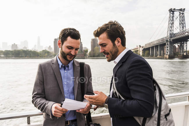 Dos hombres de negocios revisando documento en ferry en East River, Nueva York, EE.UU. - foto de stock