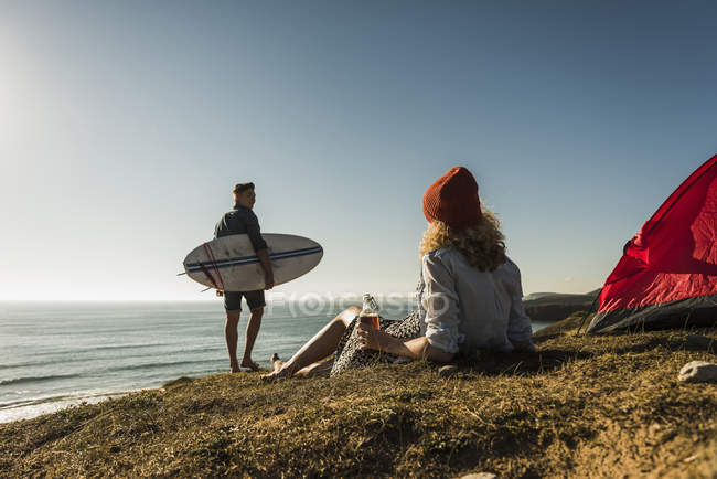 Молода пара кемпінг на узбережжі, хлопець з дошкою для серфінгу, дівчина сидить на червоному наметі — стокове фото