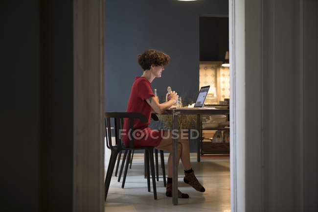 Jeune femme assise dans la cuisine et utilisant un ordinateur portable — Photo de stock