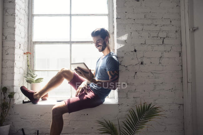 Улыбающийся молодой человек сидит на подоконнике и смотрит на планшет —  интернет, Современность - Stock Photo | #173617716
