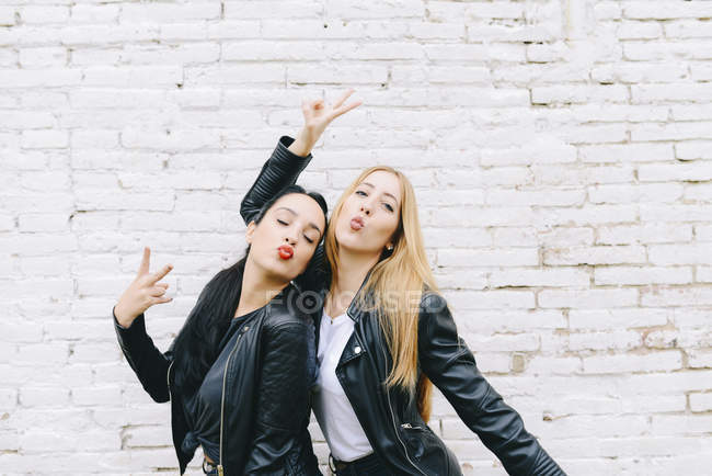 Dos mujeres jóvenes haciendo pucheros en la boca y mostrando la señal de victoria frente a la pared de ladrillo blanco - foto de stock