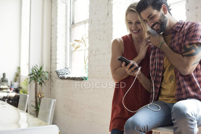 Happy young couple with cell phone at the window sharing headphones — Stock Photo