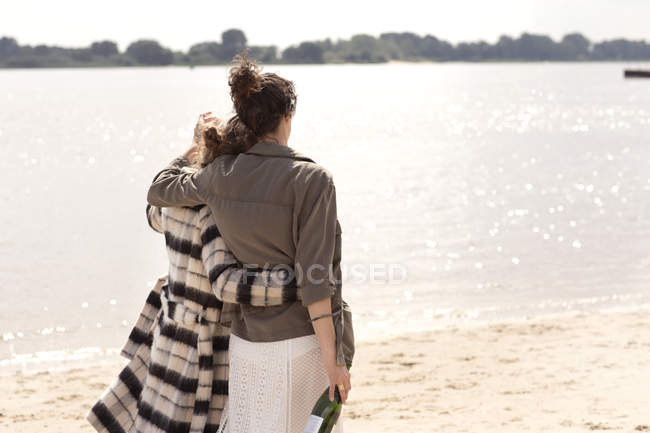 Back View Of Two Friends Walking Arm In Arm On The Beach With Bottle Of Sparkling Wine Head On Shoulder Familiarity Stock Photo