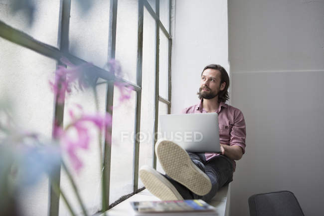 Hombre sentado en el alféizar de la ventana y utilizando el ordenador portátil - foto de stock