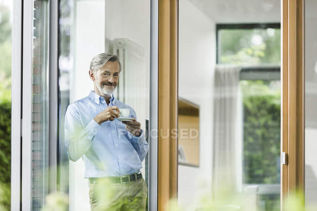 Smiling man with cup of coffee standing in his house looking through window — Stock Photo