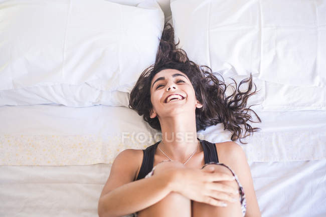 Laughing teenage girl lying on bed hugging knees — Stock Photo