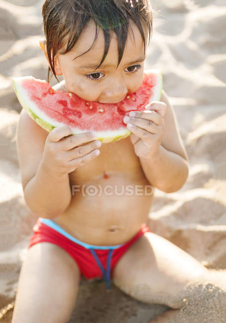 Niño sentado en la playa comiendo sandía - foto de stock