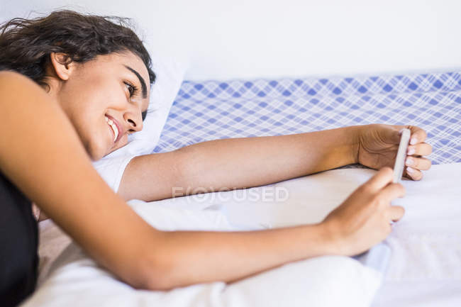 Smiling teenage girl lying on bed looking at smartphone — Stock Photo