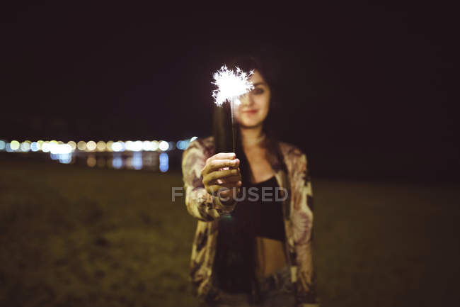 Mujer joven sosteniendo chispeante en la playa por la noche - foto de stock
