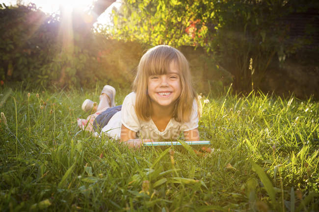 Retrato de niña sonriente acostada en el prado con la tableta - foto de stock