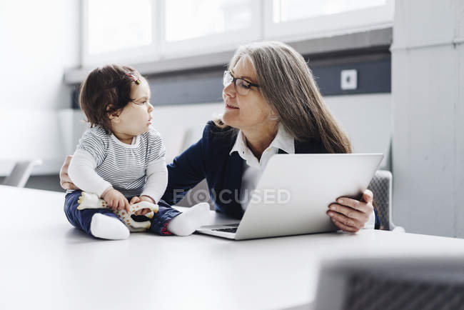 Donna d'affari anziana seduta al tavolo da conferenza con computer portatile che guarda la bambina — Foto stock