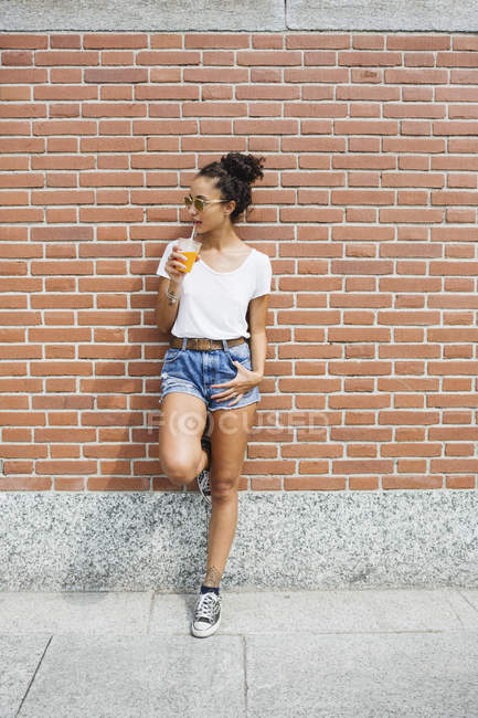 Young woman leaning against a brick wall drinking orange juice — Stock Photo