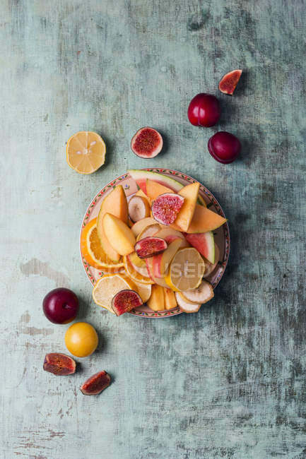 Bandeja con melón en rodajas, higo, naranja, plátano y ciruelas sobre madera gris malhumorada - foto de stock