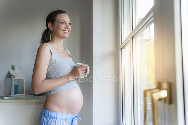 Smiling pregnant woman holding cup at window — Stock Photo