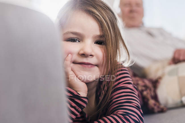 Дівчинка сидить на дивані і дивлячись на камеру з дідусь на тлі — стокове фото