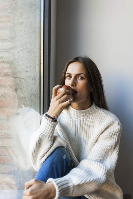 Jeune jolie femme caucasienne manger pomme à côté de la fenêtre — Photo de stock