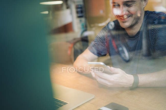 Hombre sonriente en el escritorio con teléfono celular y portátil - foto de stock