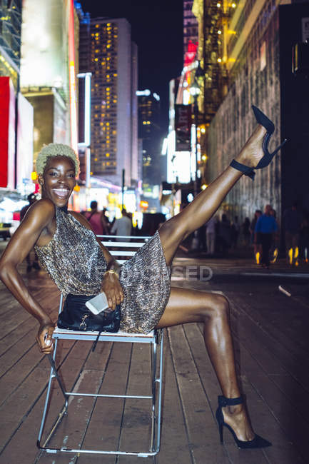 Femme américaine africaine drôle avec téléphone portable assis sur la chaise à Times Square la nuit, NY, États-Unis — Photo de stock