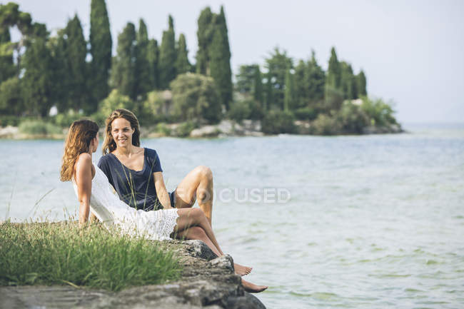 Itália, Lago de Garda, duas jovens mulheres sentadas na margem do lago — Fotografia de Stock