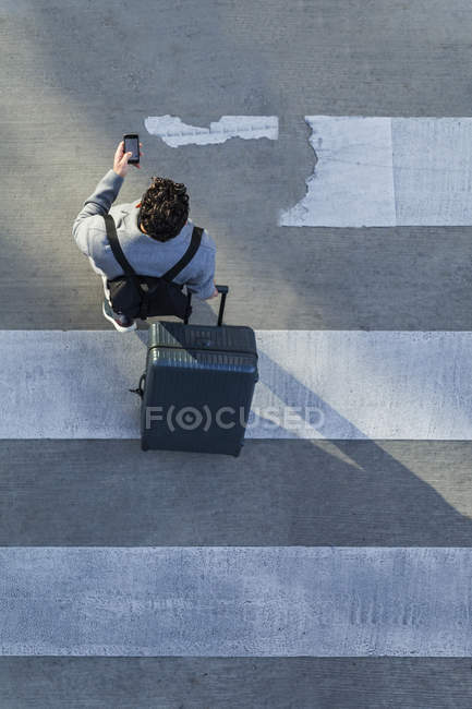 Uomo d'affari con bagaglio che attraversa la strada mentre utilizza il telefono cellulare — Foto stock