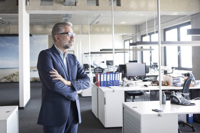 Empresario de pie en la oficina con los brazos cruzados y pensando - foto de stock