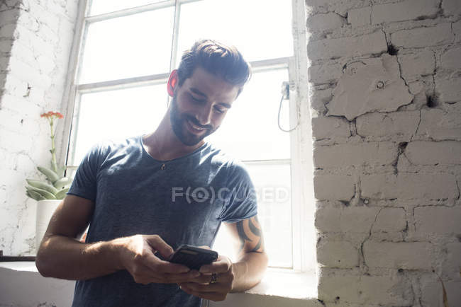 Smiling young man looking on cell phone at the window — Stock Photo