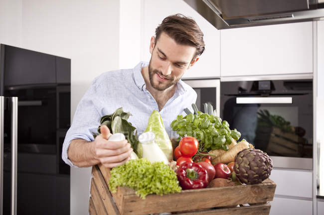Портрет молодого чоловіка з дерев'яною коробкою зі свіжих овочів і двома молочними пляшками на кухні — стокове фото