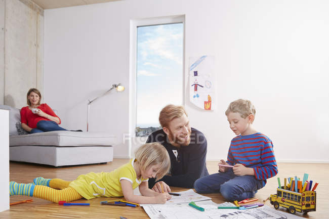 Padre acostado en el suelo con los niños pintando con la madre en el fondo - foto de stock