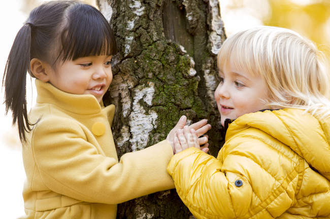 Dark-haired girl and blond boy playing next to tree trunk — affection,  idyllic - Stock Photo | #176842448
