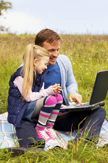 Отец и дочь сидят на лугу, отец работает над ноутбуком — стоковое фото