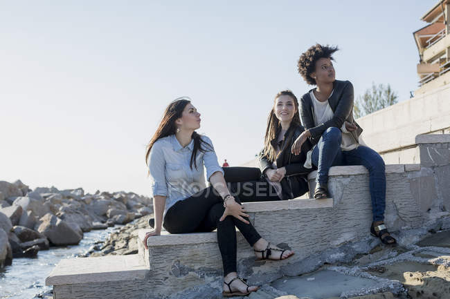 Trois jeunes femmes assises sur un mur au bord de la mer — Photo de stock