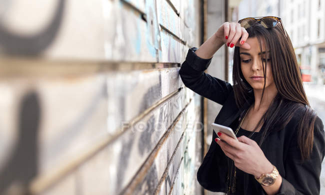 Mujer joven usando teléfono inteligente mientras se apoya en la pared en la calle - foto de stock