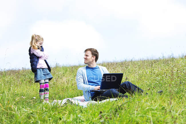 Отец и дочь сидят на лугу, ноутбук, отец смотрит на дочь — стоковое фото