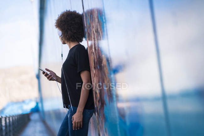Mujer joven con auriculares y teléfono inteligente apoyado contra la pared - foto de stock