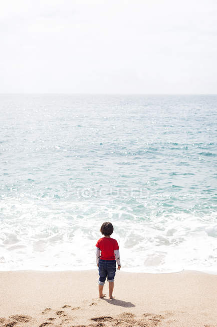 Niño pequeño de pie en la playa y mirando al mar - foto de stock