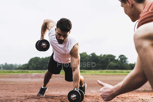 Atleta haciendo ejercicio con pesas en el campo de deportes apoyado por su compañero de entrenamiento - foto de stock