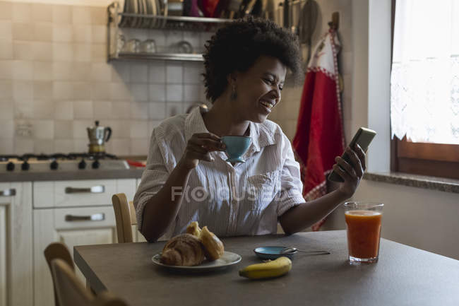 Laughing young woman with espresso cup sitting in her kitchen looking at phablet — Stock Photo