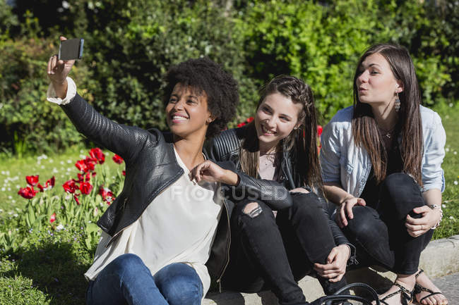Tres amigas sentadas en la hierba tomando selfie con teléfono inteligente - foto de stock