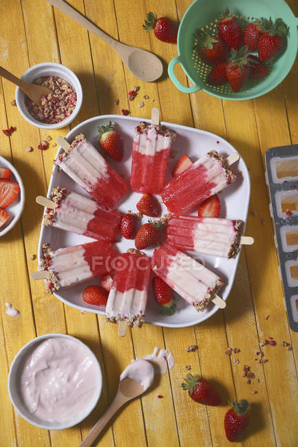 Strawberry ice lollies on platter with yogurt and cereals on yellow wooden surface — Stock Photo