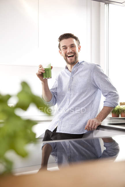 Portrait of laughing young man with green smoothie in his kitchen — Stock Photo