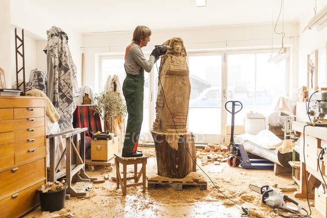 Wood carver in workshop working on sculpture with milling machine, standing on stool — Stock Photo