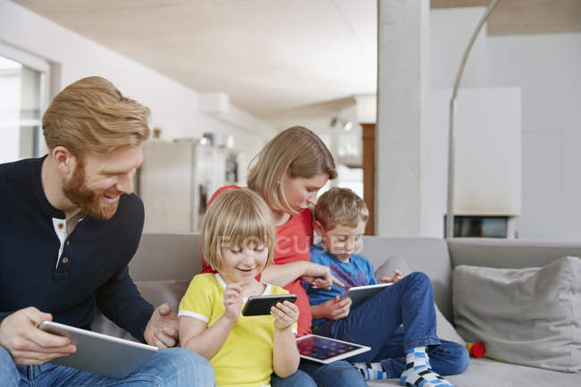 Familia que sostiene dispositivos móviles en el sofá en casa - foto de stock