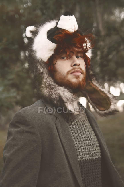 Portrait of redheaded young man wearing raccoon hat in the woods — Stock Photo