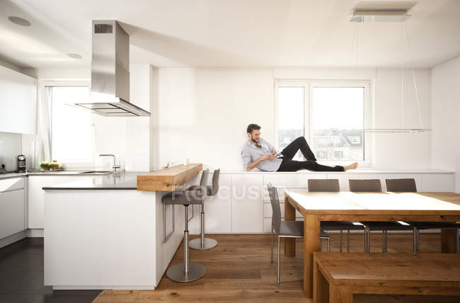 Hombre acostado en el aparador en su cocina abierta usando un teléfono inteligente - foto de stock