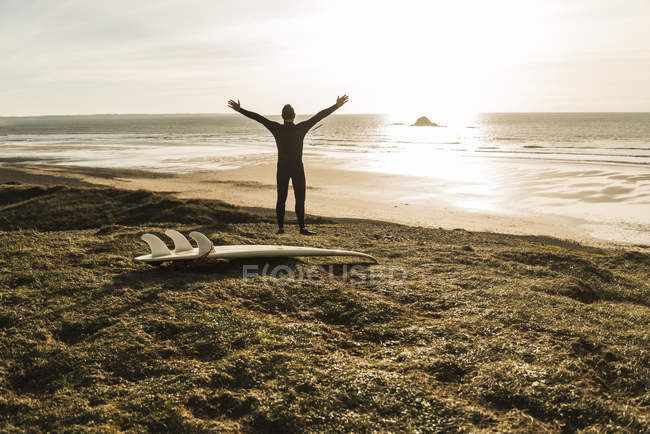 Francia, Bretagne, Finistere, Península de Crozon, hombre en la costa con los brazos extendidos y tabla de surf - foto de stock