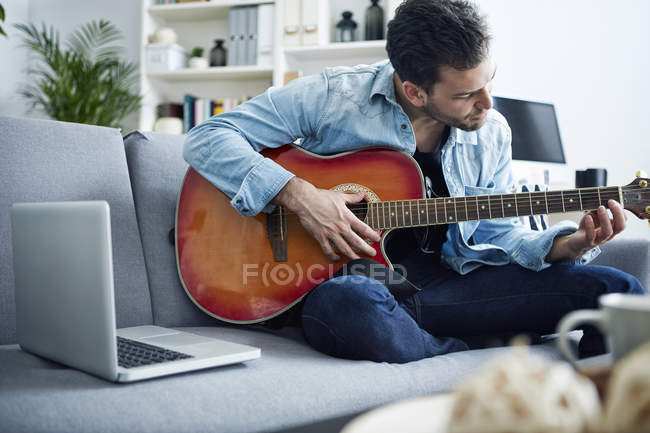 Giovane seduto sul divano a casa e suonare la chitarra accanto al computer portatile — Foto stock